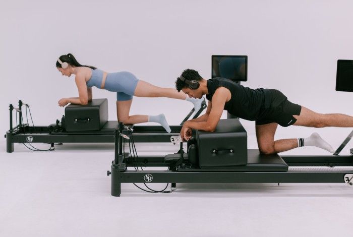 Two people, a man and a woman, are exercising on black reformer Pilates machines in a bright, minimalist studio. Both are wearing wireless headphones and athletic wear—the woman in a blue workout set and the man in black shorts and a sleeveless top with white socks. They are positioned on their hands and knees, performing a leg lift movement, each facing forward with a focused expression. Two flat screens are mounted in front of them.